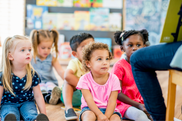 Young children seated on the floor during a library storytime program, listening to an adult reading at the front of the room.