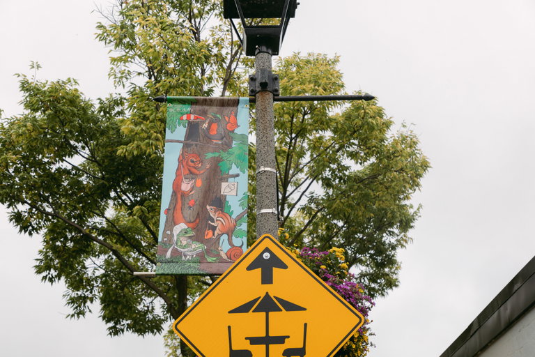 A street pole banner hanging from a metal rod. The pole banner is blue, brown, green, and orange. It illustrates squirrels shopping for acorns, a frog lounging while drinking a coffee, birds, and butterflies.