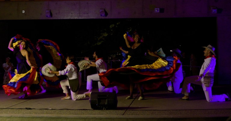 Somos Columbian Dancers performa at the Sundog Festival of Arts and Culture. The women are wearing brightly coloured traditional dresses and the men are dressed in all white with a striped hat and bag. The women are twirling in a circle and the men are down on oe knee.