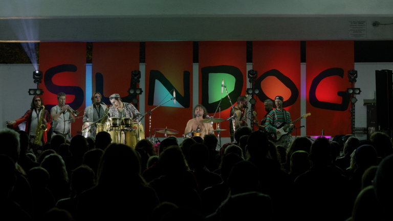 Five Alarm Funk performs on an outdoor stage at night during the Sundog Festival. The stage backdrop features large, brightly colored panels spelling "SUNDOG" in bold, abstract letters. A large crowd watches in the foreground.