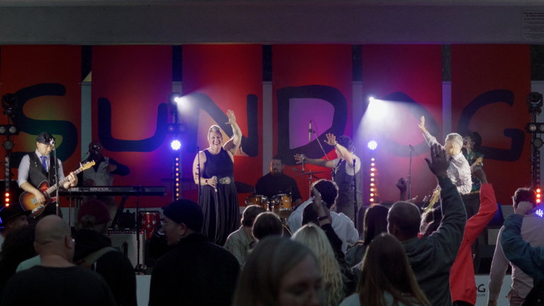 The band Queer as Funk performs on an outdoor stage at the Sundog Festival, with vibrant red panels behind them spelling "SUNDOG" in bold, artistic letters. The lead vocalist stands center stage, smiling and waving to the crowd, while other band members play guitar, drums, keyboard, saxophone, and bass. Stage lights shine brightly, and audience members dance and raise their hands, creating a joyful, energetic atmosphere.