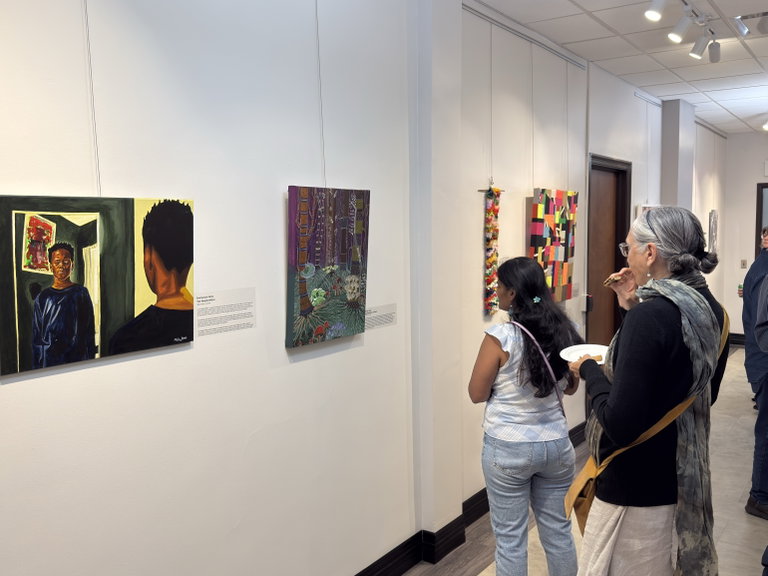 A distance shot of the exhibition hallway with visitors looking at paintings and textiles hanging on the wall.