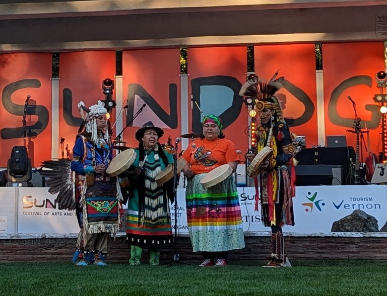 The All My Relations First Nations Dance Group performs at the Sundog Festival. The four Indigenous syilx Okanagan performers stand in traditional regalia, each holding hand drums. They are positioned in front of large red panels that spell out "SUNDOG" in bold, artistic letters.