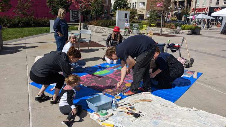 A group of people of different ages are gathered around a large painting board to create a collaborative mural with artist Monica Loney