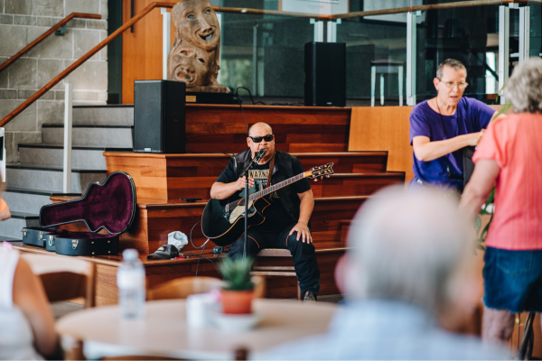 Man with a guitar sitting on steps of an atrium and speaks into a microphone as a crowd watches