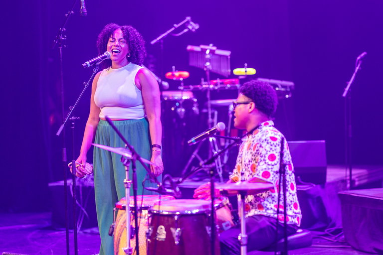 A female vocalist with curly hair singing into a microphone with a person playing hand drums seated beside her