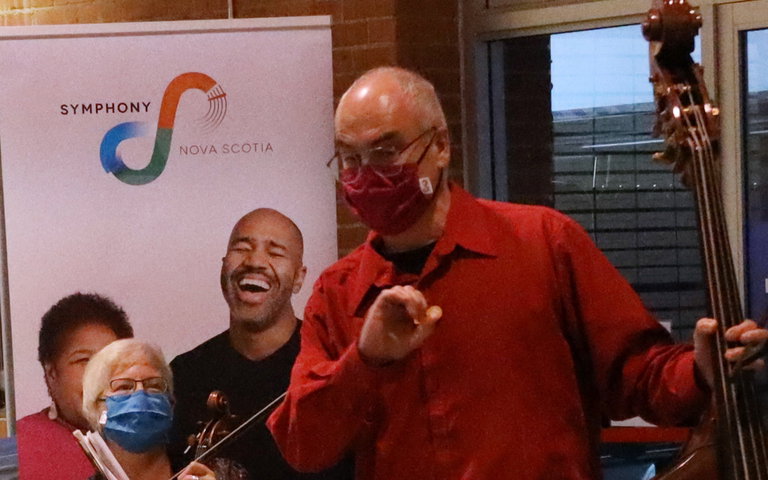 A man in a red shirt, holding a musical instrument, talks to the audience.