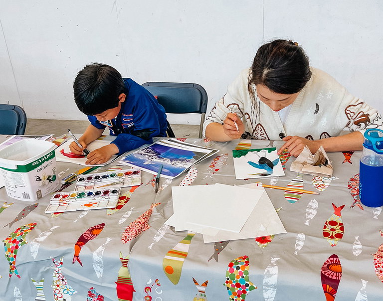 A mother and son sit side by side creating collages with a range of paper materials.