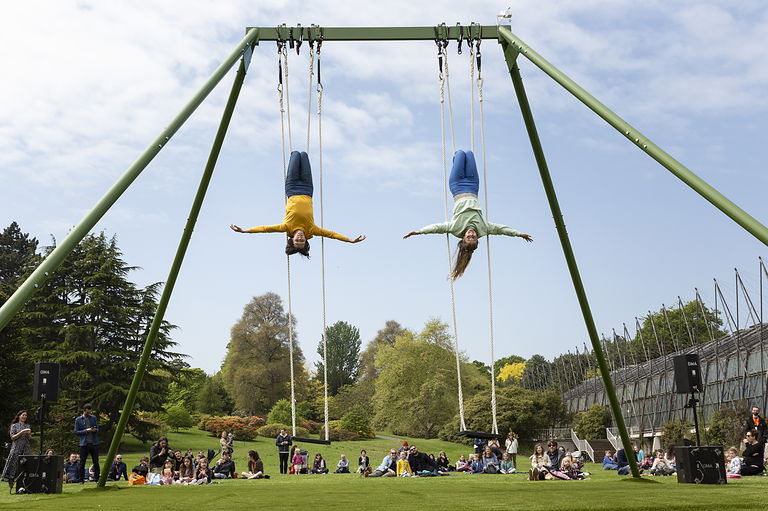 Two aerial dancers perform on a swing set in front of an outdoor crowd.