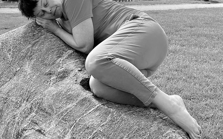 Woman curled up on a large rock with her hands cupped against the rock so as to try and listen to the rock.