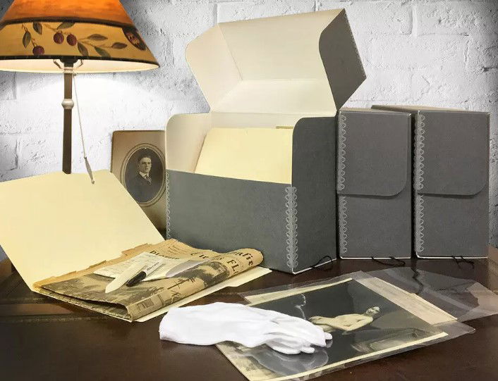 Various items on a desk, including a folder open to show a sharpie pen and newspaper inside, white cloth, images in protexctive plasict and various file holders. There is also a turn of the century photograph behind the folder