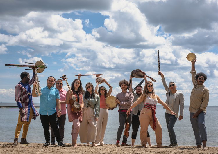 A group of 11 smiling musicians from cultures around the globe hold their instruments aloft on a beachfront.