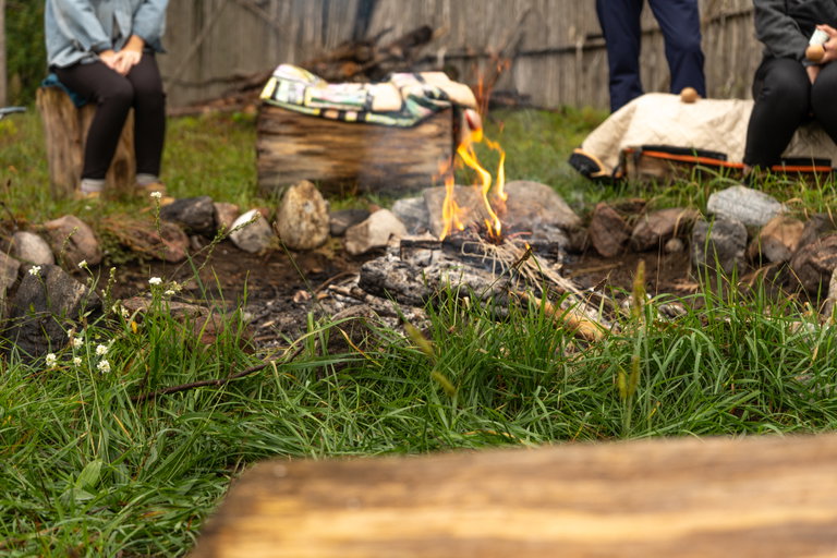 A small campfire burns within a ring of stones in a grassy outdoor setting, with flames rising from charred wood and kindling. Several people sit nearby on low stools or logs around the fire, partially visible at the edges of the scene.