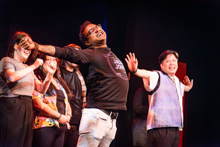 Two men are seen on improvising, extending their arms outwards like wings in an improv scene. In the background, there is a line of other improvisers cheering them on.
