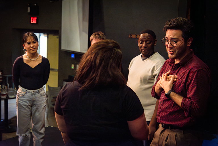 A woman in a black shirt and a man in a red shirt perform an improv scene while others watch intently.