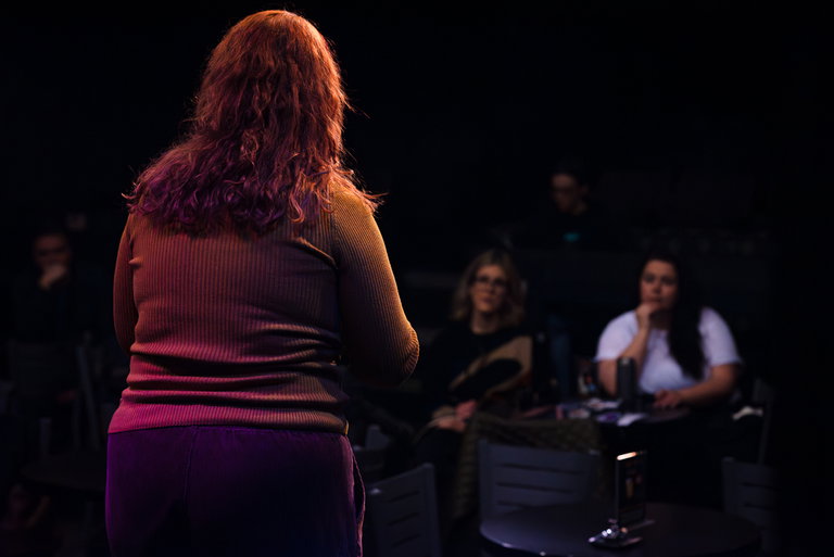 A woman in a red shirt is seen speaking to a group of people about improv comedy, which includes two women.