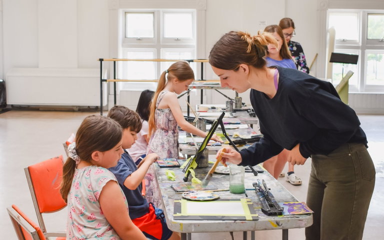 A group of youth sit at a table and paint in the Arts Centre, with a female instructor on the other side of the table.