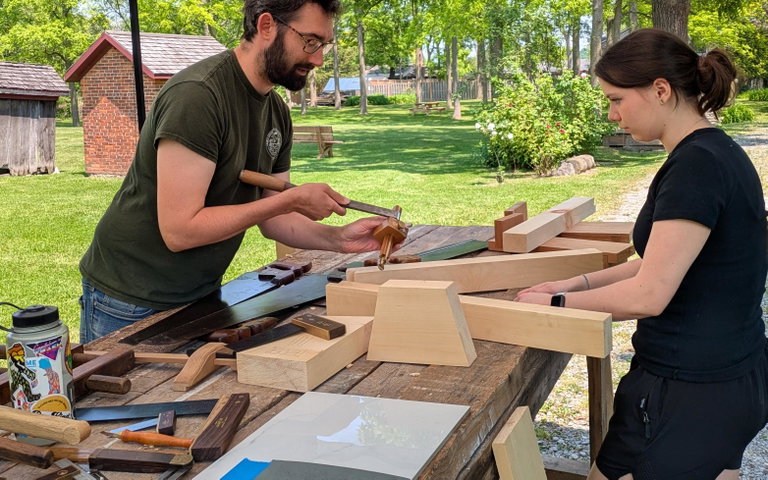 A large wooden table is scattered with various pieces of wood, saws, mallets and other tools. A man stands on one side of the table using the tools to demonstrate, while a female visitor stands on the other side of the table watching.