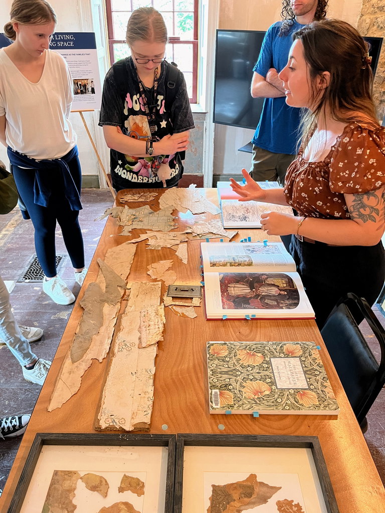 A table displaying various samples of historic wallpaper. A crowd gathers around as someone speaks.