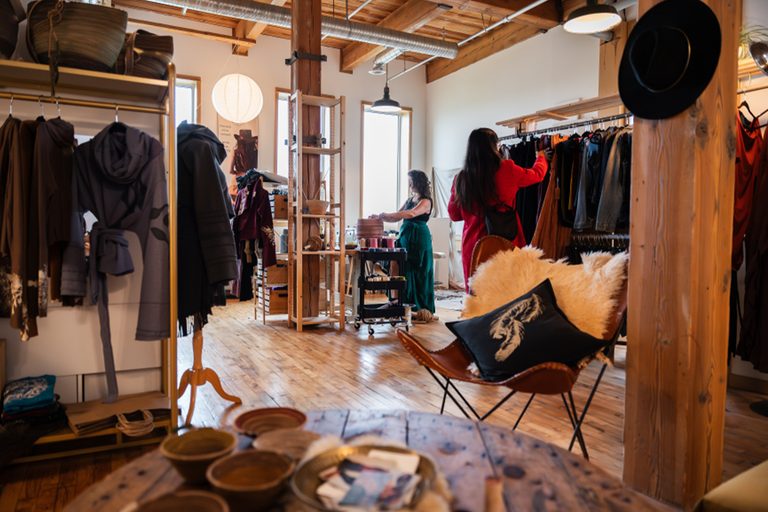 A studio of handmade clothing and baskets, with one woman browsing racks of clothes while the artist works on making a basket