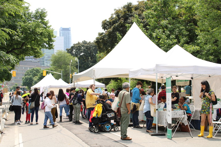 WOTS attendees walk along the white marketplace tents and shop, with Queen's Park in the background.