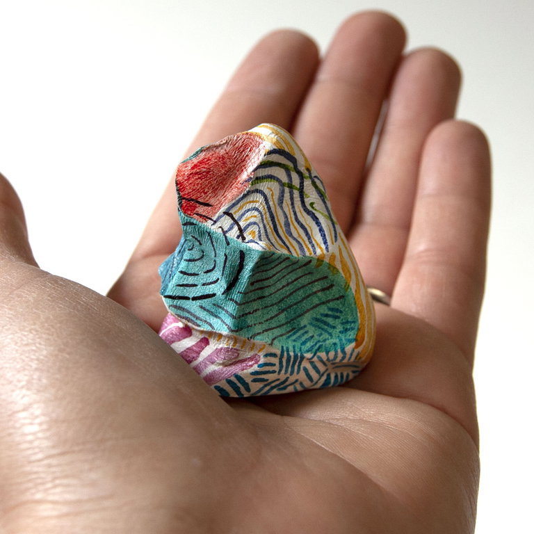 A hand holding a uniquely patterned rock. The rock has various colors and designs, including red, green, and yellow lines and shapes. The patterns appear to be hand-painted with intricate details on a white background of the stone. The image is focused on the rock with the hand serving as a scale reference, showcasing the size of the rock relative to a human hand.