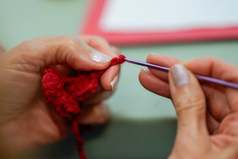 Two hands working with a knitting needle and red yarn.