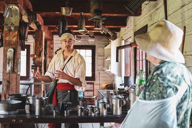A young man is in character as a Tinsmith in 1815 and surrounded by tinware in a historic building as he speaks to a visitor