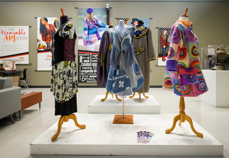 Three dresses on displayed on mannequins. The dress on the left is black and purple with clocks printed on a light coloured fabric. The dress in the centre is stitched together of striped, dotted, and floral blue fabrics. The dress on the right has long sleeves, with circular patches sewed in a painterly fashion, of purple, pinks, oranges and greens.