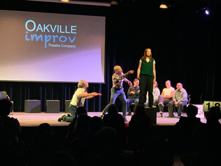 Oakville Improv performers standing on stage. One performer is on top of a black box, standing and facing the audience.