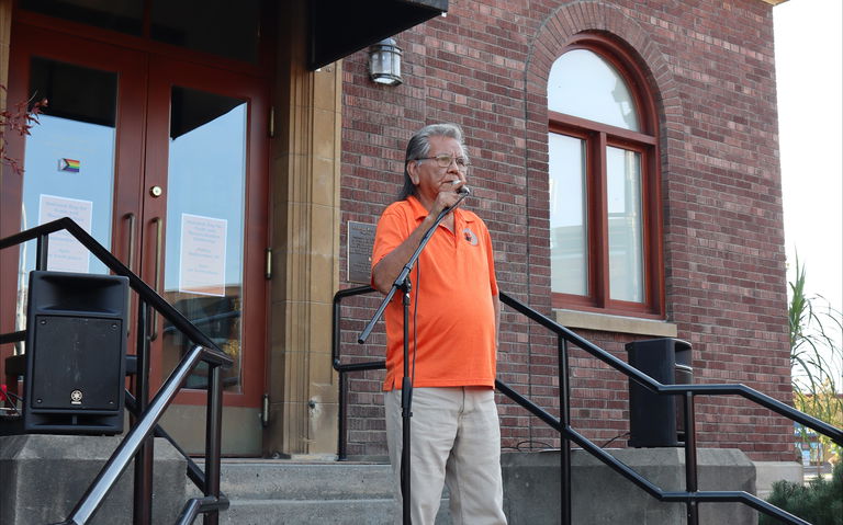 Knowledge Sharer Louis Thomas standing on the steps of a brick building and speaking into a microphone. He is wearing tan pants, black shoes, aviator style framed glasses and an orange shirt polo style shirt with an emblem that has a black handprint and a grey crescent over his chest on the left side.