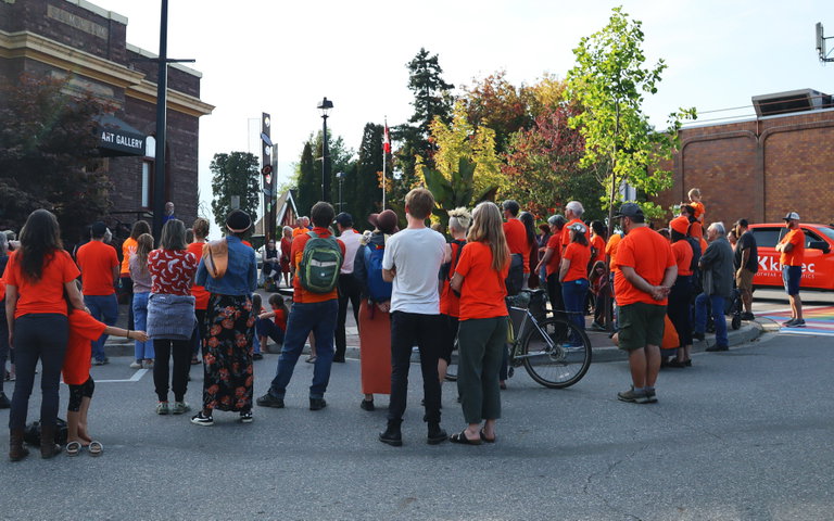 Outside a brick building with an awning that reads "Art Gallery" a group of approximately 80 people wearing orange shirts are gathered.