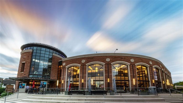 A wide-angle view of The Rose Brampton theatre, showcasing its modern curved brick-and-glass facade under a dramatic, fast-moving sky. Large arched windows glow warmly from within, and outdoor patio seating with umbrellas lines the front steps. The photo captures the grandeur and architectural elegance of the venue at dusk.  Let me know if you'd like a shorter or AODA-compliant version for web or social media!