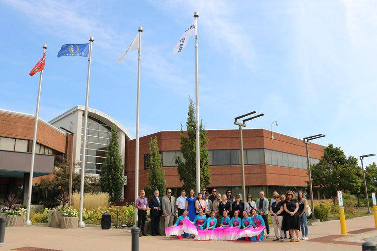 A group of Town staff, community members, and the Oriental Arts Club performers gathered together in front of Town Hall in Oakville. There are four flag poles behind them, one with a white and black Culture Days flag raised to the top.
