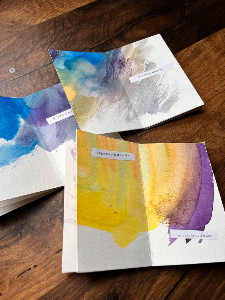 Three hand-painted paper booklets with cut-out poetry excerpts laid on a brown, wooden table.