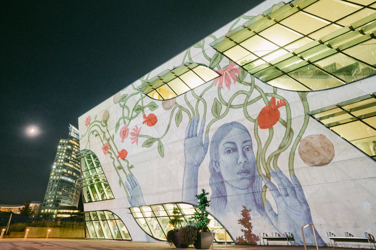 Video artwork featuring a blue human with green vines coming out of their fingers, pink and red flowers and pomegranites,projected on a building.