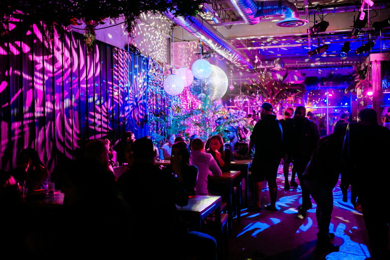 Full taproom featuring tropical lights in pink and blue, in the background is the Reflektieren area.
