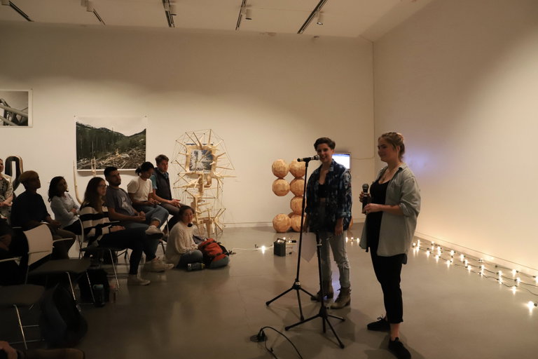 Two people on right side with microphone in hand, in front of small seated crowd on the left. Art including instillations behind crowd.