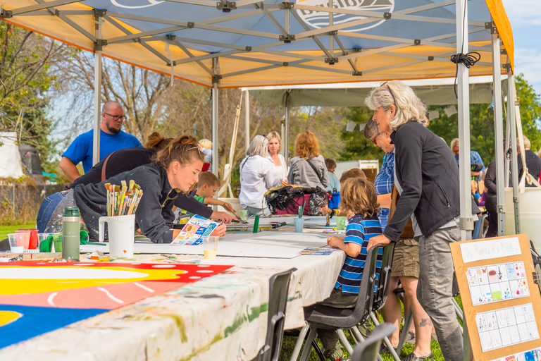 People sitting and standing around a table under a tent painting mural tiles.
