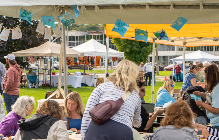 A group of people sitting around a table under a tent painting mural tiles.