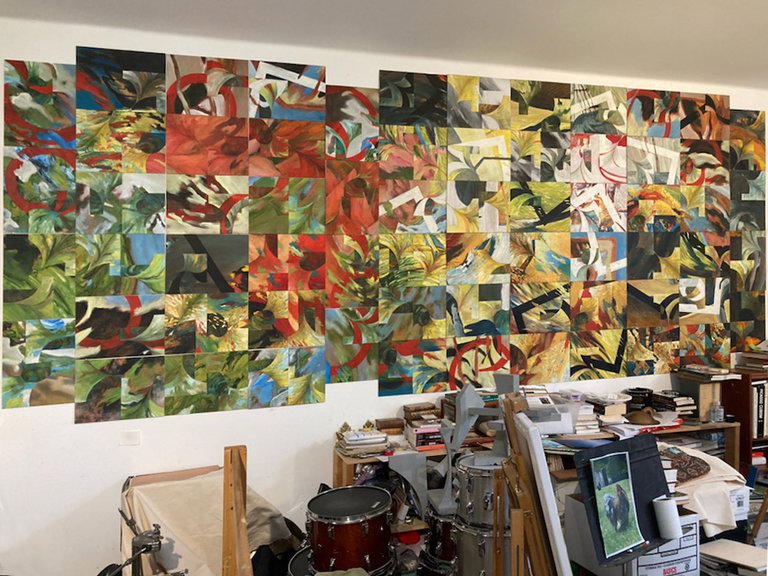 In-studio view of wall-sized abstract painting comprising many smaller paintings of various colours including reds, greens, and yellows; curved and funnel-shaped forms are interspersed and entangled with geometric shapes and lines with harder edges