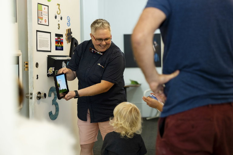 An artist smiles at a small child while holding up a tablet in a studio space