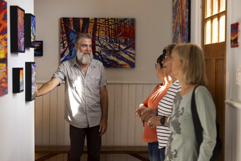 Four people stand in a gallery studio setting surrounded by brightly coloured landscape paintings hanging on the walls around them