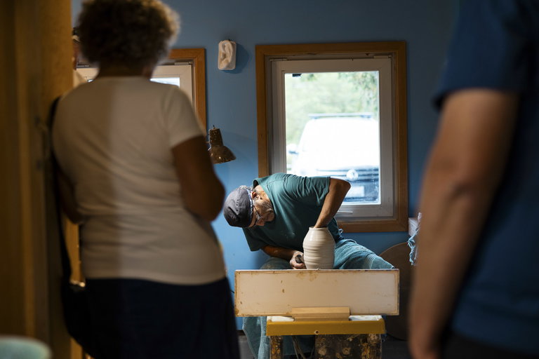 A person throws a clay pot on a potter's wheel in a studio space. In the foreground there are two people watching the deminstration.