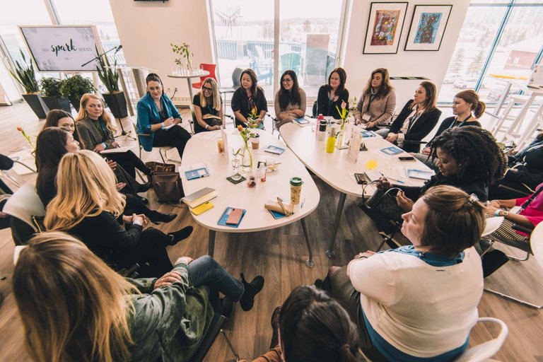 The image displays the Sandbox Centre's interior space and how it is used. People are gathered around tables in the middle of the room for conversation. It is a bright space with lots of windows and art displayed.