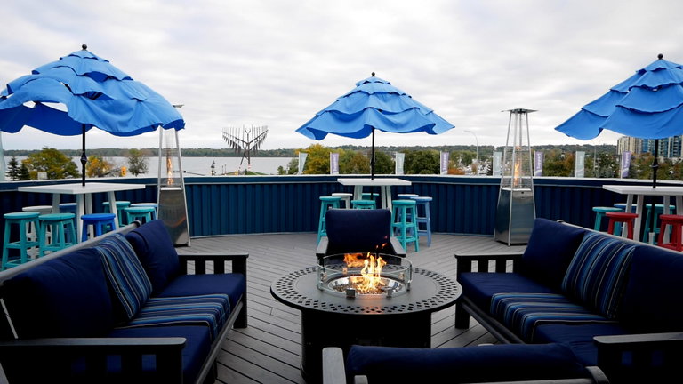 The image displays the Sandbox Centre's exterior space. It is a rooftop patio overlooking Barrie's waterfront, and the iconic, 72-feet tall Spirit Catcher statue. The patio includes modern tables with colorful umbrellas and chairs, as well as outdoor heaters, and couches around a campfire in the centre.