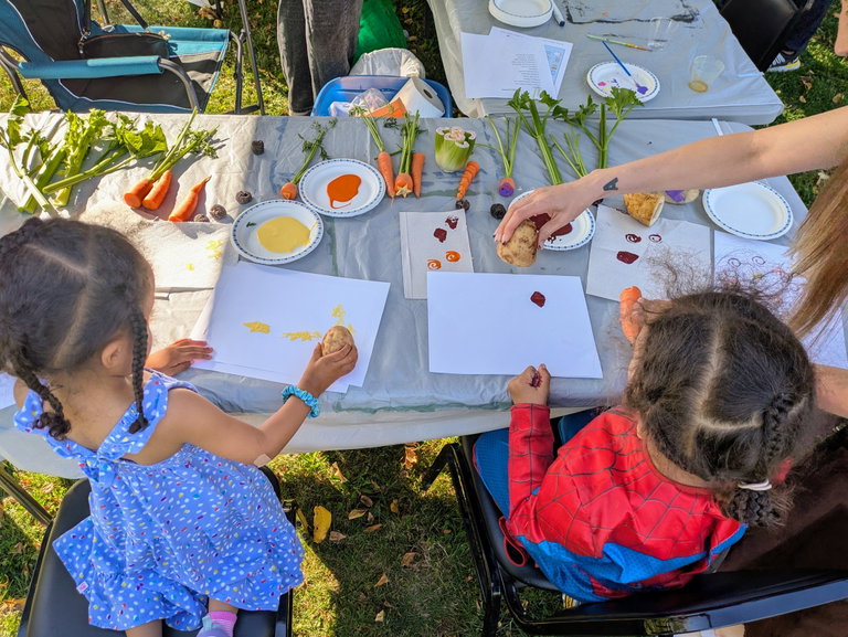 Children stand over a table, using cut carrots, celery and potatoes to stamp paint onto paper.