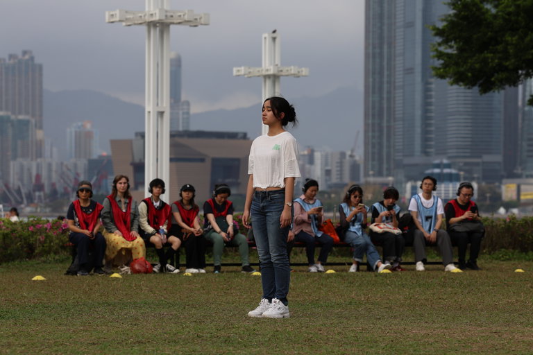 This photo shows the participants of Playing Fields. Standing alone on a soccer field is a young woman, with her arms by her sides and her eyes closed. Behind her, 10 participants of the program can be seen sitting on a bench, wearing red and blue jerseys and headphones.
