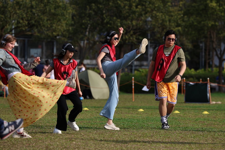 This photo is of 4 people participating in Playing Fields. In the centre, a woman is wearing jeans and is kicking her foot high in the air. All of the participants are standing on a green soccer fields and are wearing red jerseys and headphones.