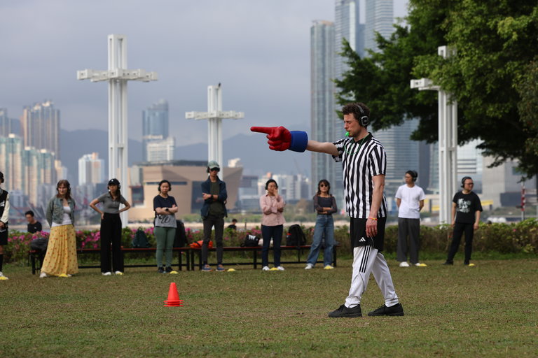 This photo is of the participants of Playing Fields. Clearly seen in the photo is a referee, complete with a black and white striped shirt, a green whistle and a large red glove. He is pointing ahead of him with the gloved hand and has the whistle in his mouth. Behind him, there are 10 participants of the program, each wearing headphones.